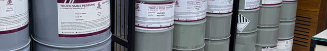 A retail display of industrial paint or coating products, featuring large gray metal drums and smaller white bottles with gold caps arranged on black metal shelving units over a light marble floor.